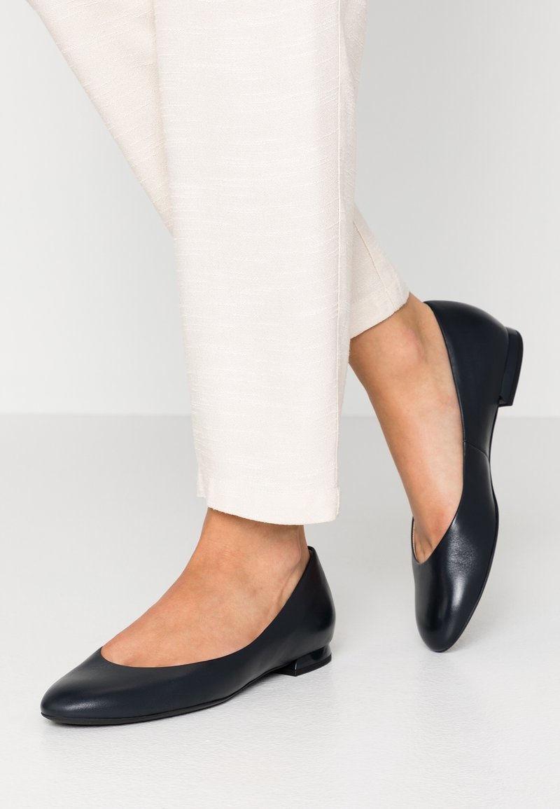 Ballerine in pelle nera con punta affilata, tacco basso e aperture laterali, abbinate a pantaloni eleganti di colore chiaro. Texture liscia.