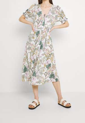 Femme portant une robe midi blanche avec un imprimé floral coloré, des manches courtes bouffantes et des sandales compensées beige, debout devant un fond uni.