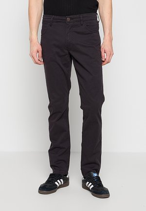 Mężczyzna ubrany w czarne dopasowane spodnie i czarne buty sportowe Adidas z białymi paskami, stojący na jednolitym jasnoszarym tle.