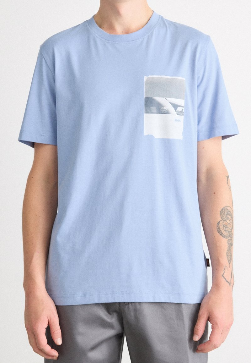 T-shirt bleu clair à manches courtes avec une petite impression photo en niveaux de gris sur la poitrine gauche, porté par une personne avec un tatouage de serpent sur l'avant-bras droit.