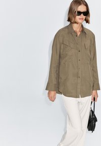 Khaki button-up shirt with two chest pockets, long sleeves, and a relaxed fit, paired with white pants and a small black handbag.