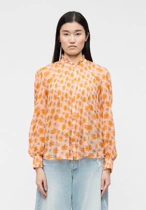 Mujer vestida con una blusa rosa de manga larga, con volantes y botones, estampada con flores naranjas, combinada con vaqueros azules claros desgastados.