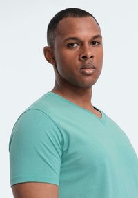 Mladý muž s krátkými vlasy, oblečený v světle zeleném tričku s výstřihem do V, se sebevědomě dívá přes rameno na jednobarevném pozadí.