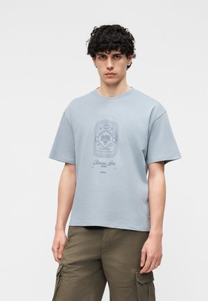 Ung mand med krøllet mørkt hår iført lyseblå t-shirt med grafik og olivengrønne cargobukser mod en ensfarvet baggrund.