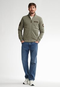 Grüne Zip-Jacke aus weichem Stoff, mit Schulterbesätzen und hohem Kragen, kombiniert mit blauen Jeans und weißen Sneakern.