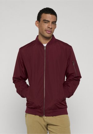 Hombre con cabello rizado corto que lleva una chaqueta bomber burdeos con cremallera y pantalones beige, con las manos en los bolsillos de la chaqueta, de pie contra un fondo liso.