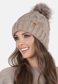 Cappello di lana lavorato a maglia beige con una texture a costine e un morbido pompon in cima. Presenta un'etichetta in pelle marrone sul lato.