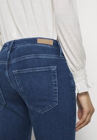 Mörkblå denimjeans med en slät yta, med bakfickor och en tan-färgad läderetikett vid midjan. Vit långärmad blus synlig.