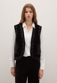 Gilet di velluto nero con intricate decorazioni ricamate lungo i bordi, indossato sopra una camicia bianca, caratterizzato da un design senza maniche e una texture liscia.
