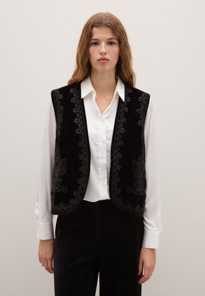 Chaleco de terciopelo negro con intrincados patrones bordados a lo largo de los bordes, llevado sobre una camisa blanca, con un diseño sin mangas y una textura suave.