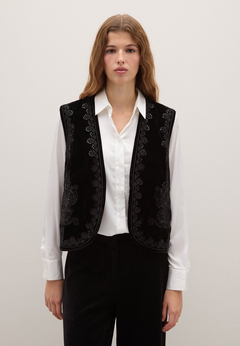 Gilet di velluto nero con intricate decorazioni ricamate lungo i bordi, indossato sopra una camicia bianca, caratterizzato da un design senza maniche e una texture liscia.