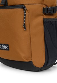 Mochila marrón y negra con material texturizado, que cuenta con un bolsillo lateral, una cremallera negra y una etiqueta con el logo. El tirador de la cremallera tiene un diseño anudado.