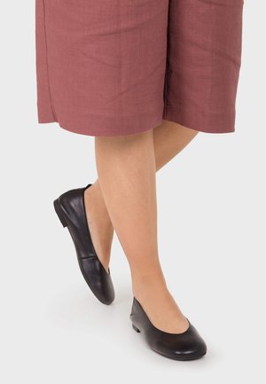 Piernas cruzadas con zapatos de cuero negros de tacón bajo y una falda burdeos hasta la rodilla sobre un fondo claro y liso.