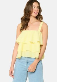 Rumena gingham bluza z večplastnimi volančki, tankimi naramnicami in ohlapnim krojem, v kombinaciji s svetlo modrimi visokim pasom kavbojkami. Teksture tkanine.