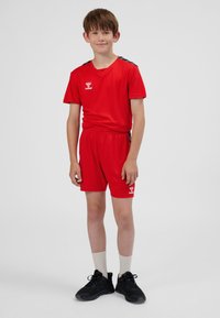 Rood sportshirt met korte broek, met logo op de borst en strepen aan de zijkant. Het materiaal lijkt lichtgewicht; het model draagt zwarte schoenen en witte sokken.