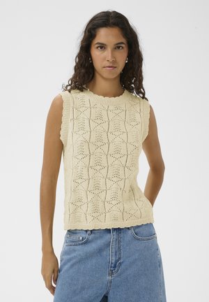 Młoda kobieta o kręconych włosach nosi kremowy, bezrękawnik z dzianiny z wzorami oraz niebieskie, wysokowalne dżinsy, stoi na tle jednolitego tła.