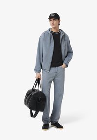 Giacca di jeans e pantaloni in blu chiaro, t-shirt nera, cappellino nero con logo, mentre tiene una borsa da viaggio nera, in piedi su uno sfondo bianco.