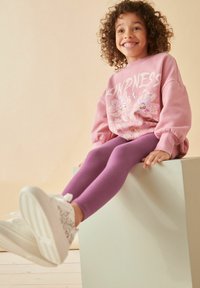 Rosa sweatshirt med texten "Vänlighet" och blommönster, ihop med lila leggings och vita sneakers med ett strukturerat mönster.