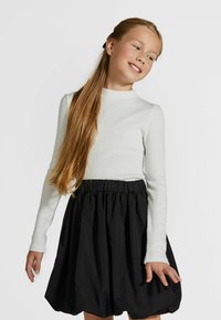 Młoda dziewczyna z długimi blond włosami, ubrana w białą bluzkę z długim rękawem i czarną spódnicę balonową, uśmiechająca się i patrząca na bok.