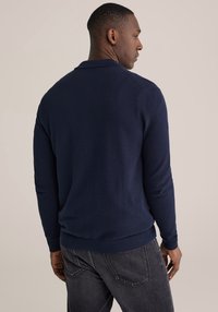 Tmavomodrý pletený sveter s textúrovaným povrchom, vybavený klasickým golierom, rebrovanými manžetami a lemom, v kombinácii s tmavými džínmi.