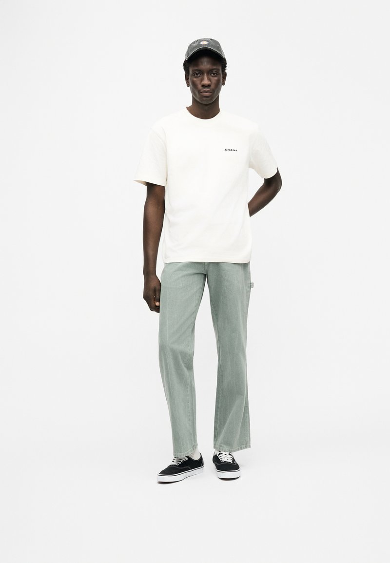 Licht crèmekleurig t-shirt met een klein zwart logo, gecombineerd met lichtgroene wijde broekspijpen en zwarte sneakers. De pet is donkergrijs met een logo.