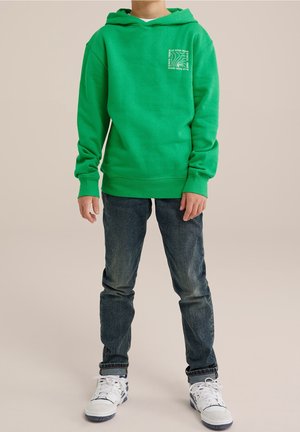 Groene hoodie met een kleine witte afbeelding op de linkerborst, gecombineerd met donkerblauwe jeans en witte sneakers. Glad materiaal, ontspannen pasvorm.