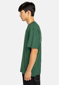 T-shirt verde in cotone con una vestibilità rilassata, maniche corte e scollatura tonda. Presenta un design stampato sul retro e un branding sottile sul davanti.