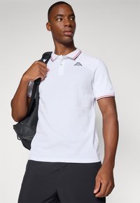 Hvit kortermet polo skjorte med rød og marineblå stripete krage, prydet med en svart logo på venstre bryst, sammen med mørke shorts og en svart bag.