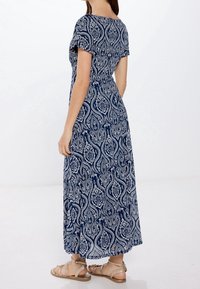 Robe maxi bleu marine avec motif cachemire blanc, manches courtes et design fluide. Le mannequin porte des sandales beige clair.