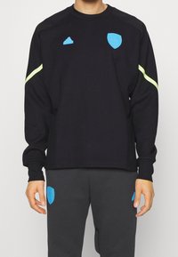 Felpa nera con accenti blu e gialli, dotata di scollo rotondo e polsini a costine. Abbinata a pantaloni della tuta grigio scuro con dettagli del logo blu.