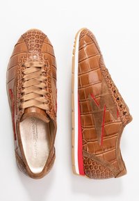 Bruna sneakers med krokodilstruktur, med detaljer i mocka och en röd blixtdesign. Platta gummisulor med vit och röd detaljer.