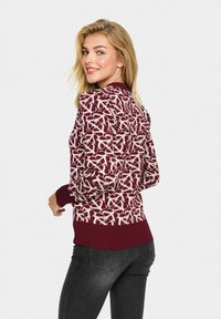 Pull marron à motifs avec des designs blancs entrelacés. Coudières et ourlet côtelés. Porté avec un jean foncé, la vue arrière affiche le design et l'ajustement.