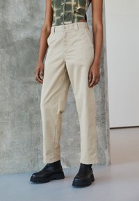 Pantalons beiges taille haute en toile de coton, avec une coupe droite et une fermeture à bouton sur le devant, associés à des bottes noires volumineuses.