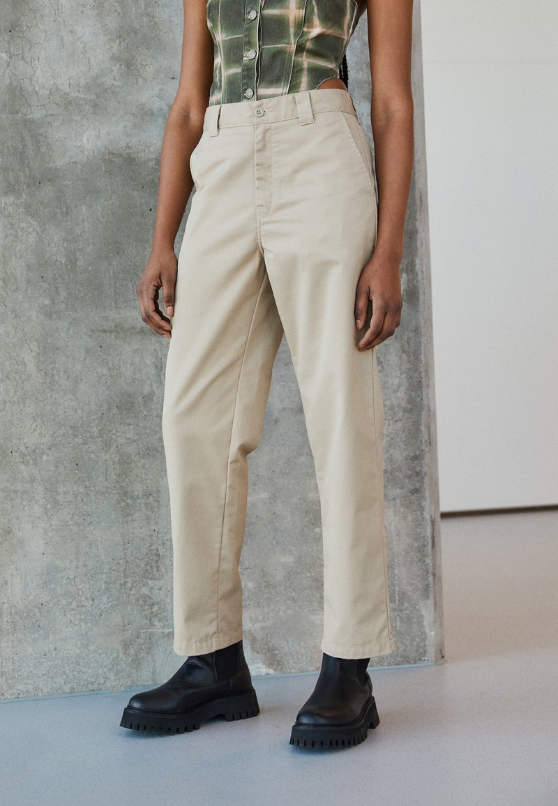 Pantalons beiges taille haute en toile de coton, avec une coupe droite et une fermeture à bouton sur le devant, associés à des bottes noires volumineuses.