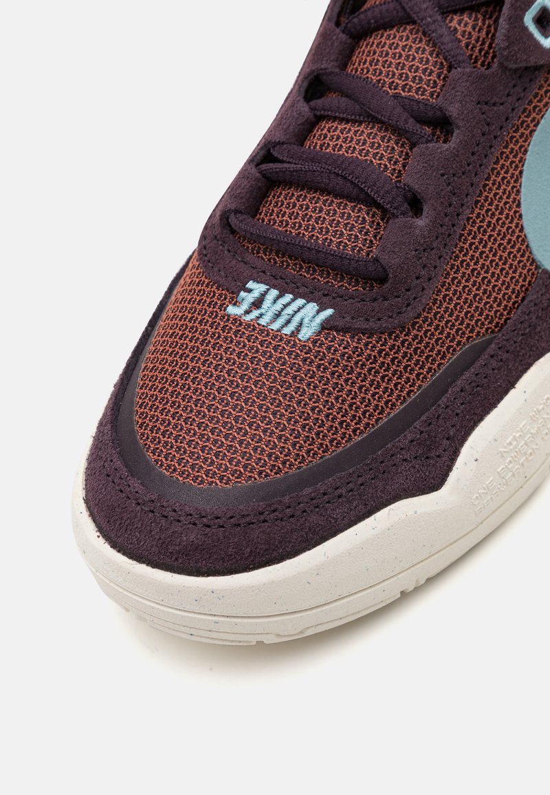 Sneaker Nike avec une tige en maille marron, accents en suède violet foncé, marquage en teal et une semelle intermédiaire blanche texturée avec des éclats.