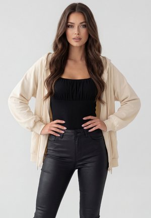 Frau mit langen, welligen braunen Haaren, die einen beigen Reißverschluss-Hoodie, ein schwarzes ärmelloses Oberteil und schwarze Lederhosen trägt und mit den Händen in der Hüfte steht.