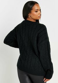 Mustan pitkän neuleen, jossa on kuvioitu pinta, ylisuuri malli ja ribbaushelma. Pitkät hihat ja pyörökaula-aukko korostavat kodikasta muotoilua.