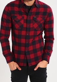 Czerwona i czarna koszula flanelowa w kratę, z dwoma kieszeniami na piersi, zapięciem na guziki z przodu oraz długimi rękawami. Posiada zaokrąglony dolny brzeg i miękką teksturę.