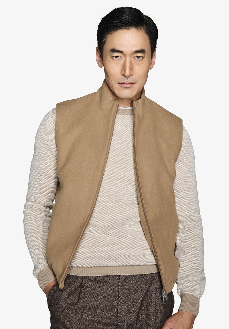 Tan ærmeløs vest med høj krave, lavet af et blødt stof, over en lys mønstret sweater. Vestene har en frontlynlås og sidelommer.
