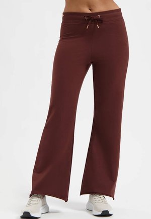 Femme portant un pantalon marron à jambes larges avec taille à cordon et baskets blanches, debout devant un fond uni.