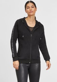 Black zip-up hoodie with a textured finish, featuring a hood, side pockets, and white branding along the sleeves. Paired with shiny leggings.