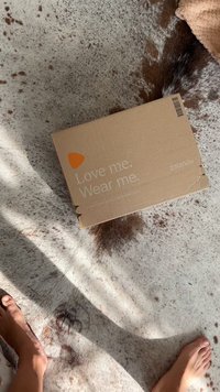 Caixa de papelão com um exterior castanho, apresentando um logotipo laranja brilhante e o texto "Ame-me. Use-me." na parte de cima, colocada sobre uma superfície texturizada.