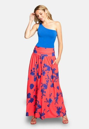 Žena v modrom jednoramennom topu a dlhej koralovej sukni s modrým kvetinovým vzorom, stojaca s jednou rukou pri vlasoch a druhou v vrecku.