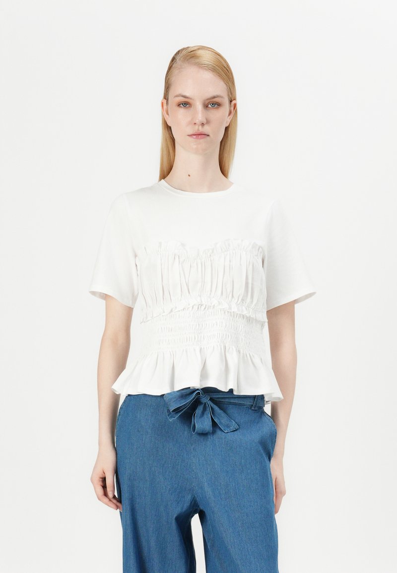 Haut blanc à manches courtes avec texture froncée et ourlet en volants, associé à un pantalon en denim bleu à jambes larges avec une ceinture nouée.