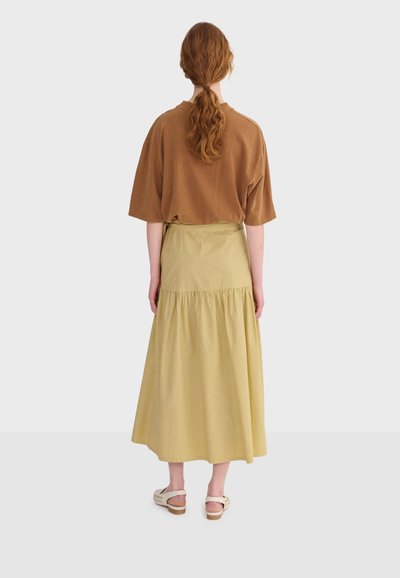 Žena s crvenom kosom u repu, odjevena u smeđu kratku majicu, žutu midi suknju i bijele sandale, stoji okrenuta leđima na bijeloj pozadini.