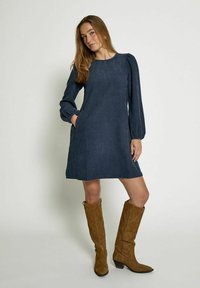 Navyblaues Kleid mit langen Puffärmeln, knielangem Saum und seitlichen Taschen. Der Stoff hat eine strukturierte Oberfläche. Braune Wildleder-Stiefel bis zum Knie.