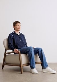 Felpa con zip blu navy e colletto a righe bianche, jeans in denim chiaro e sneakers bianche; seduto su una sedia con telaio in legno e rivestimento beige.