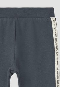Donkergrijze joggers met een geribde tailleband. De zijkanten hebben strepen met de tekst "CLUB CUTENESS" in crèmekleur. Zachte, elastische stof.