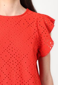 Camicetta rossa in tessuto traforato, con motivo a diamante e maniche corte arricciate. Texture liscia, scollatura a girocollo e buchi aperti su tutta la superficie.