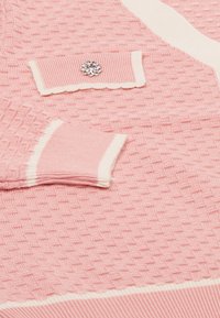 Rózsaszín kötött pulóver texturált mintával, fehér díszítéssel, bordás mandzsettával és dekoratív kristályvirág részlettel a zsebében.